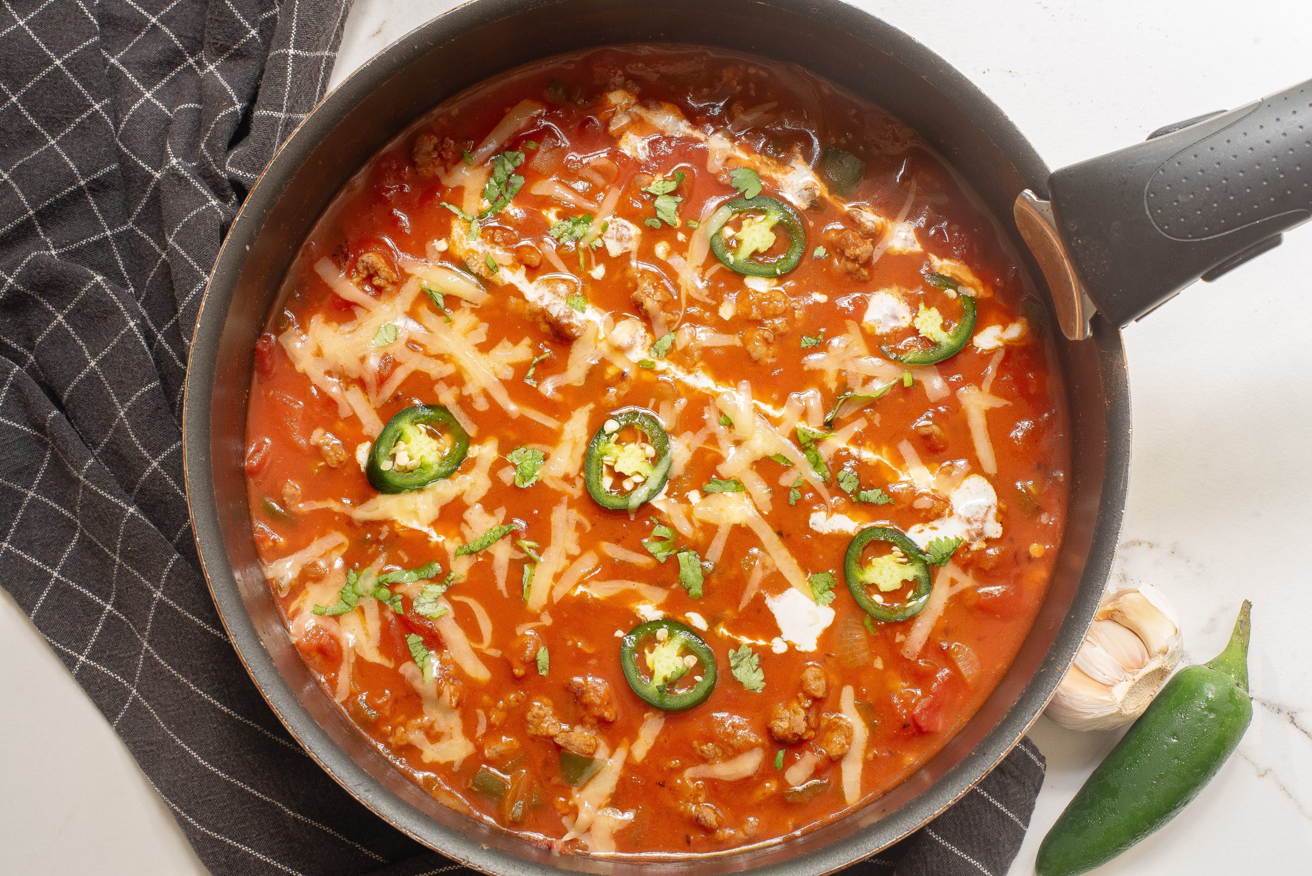 A pan of Keto chili garnished with sliced jalapeños, shredded cheese, and cilantro, placed on a black checkered cloth with garlic and a chili pepper beside it.