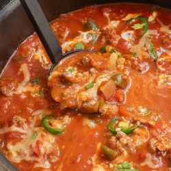 A pot of tomato-based soup with chunks of meat, sliced jalapeños, melted cheese, and vegetables, being stirred with a ladle.