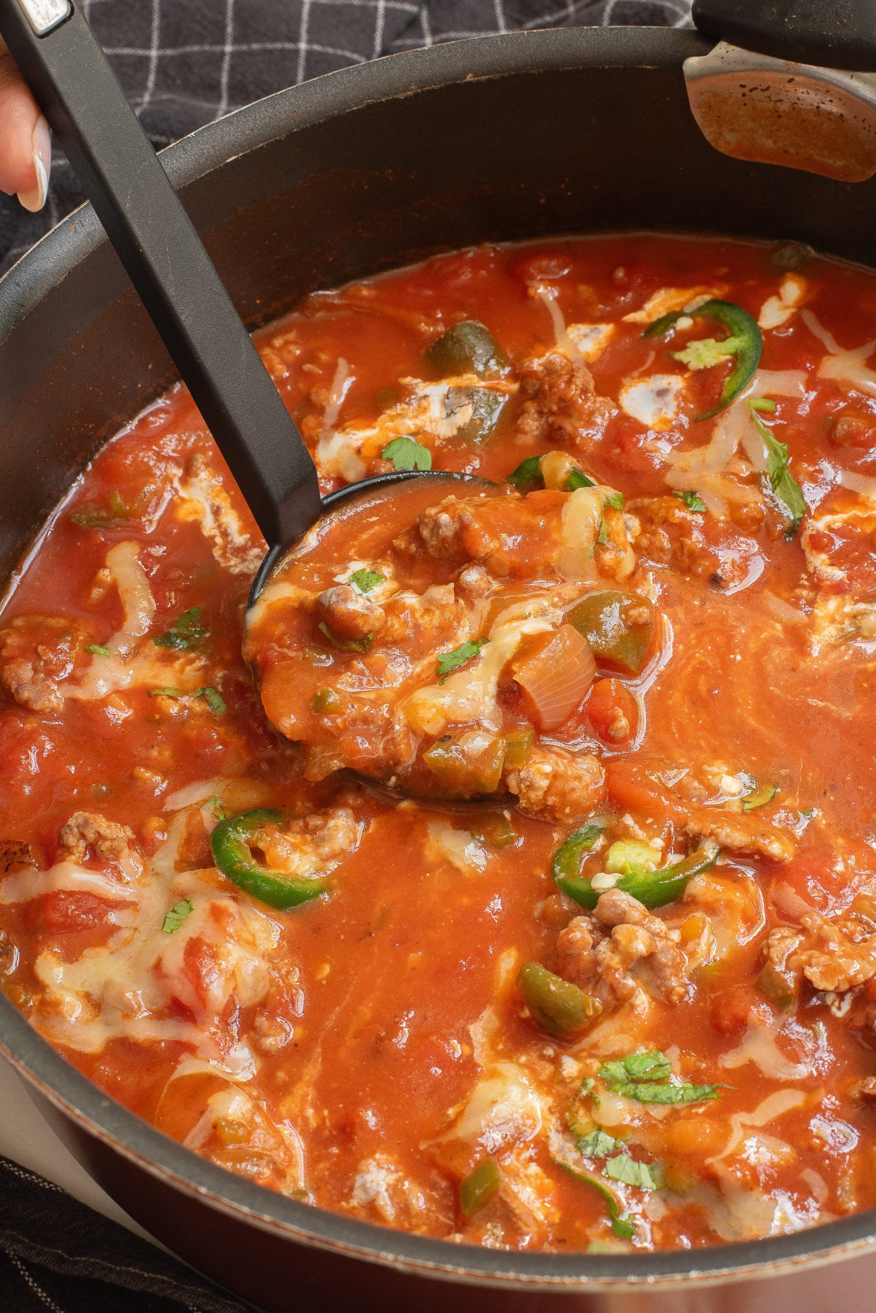 A pot of tomato-based soup with chunks of meat, sliced jalapeños, melted cheese, and vegetables, being stirred with a ladle. It's an easy low carb recipe.