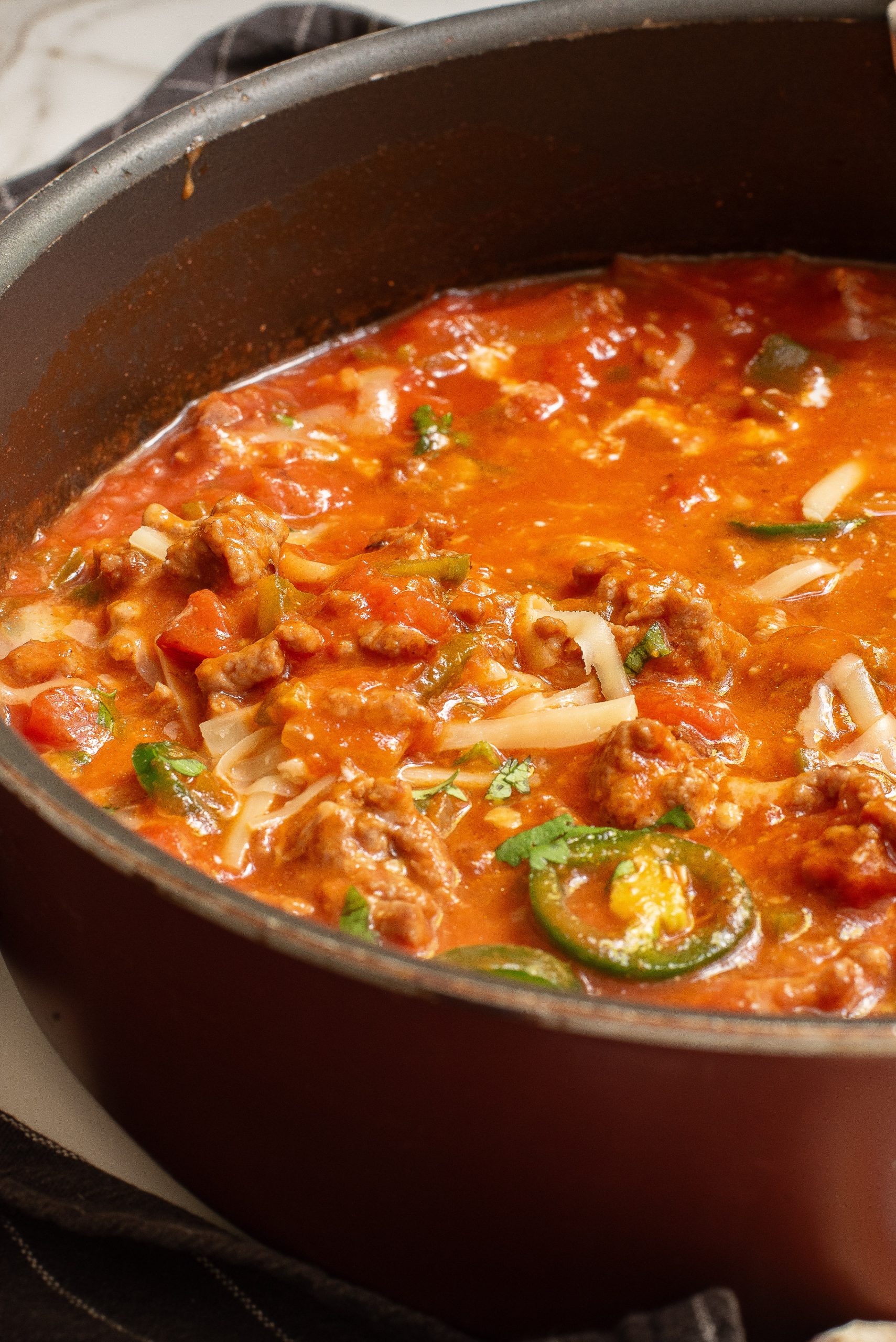 Close-up of a pot filled with a chili containing meat, green peppers, jalapeño slices, and garnished with shredded cheese.