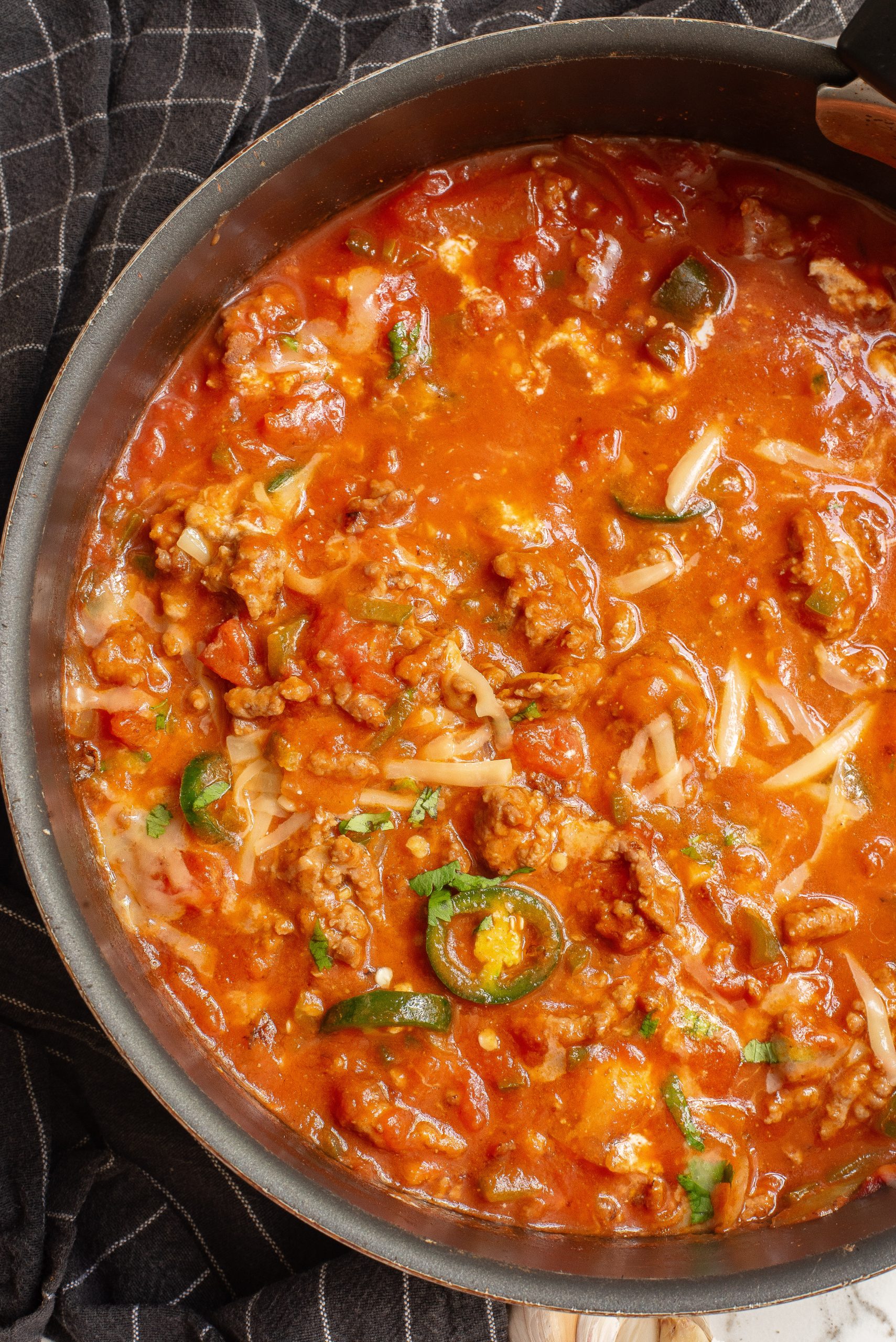 A bowl of spicy soup with sausage, peppers, and cream sits on a table next to a pot of soup, a jalapeño, cloves of garlic, and cilantro.