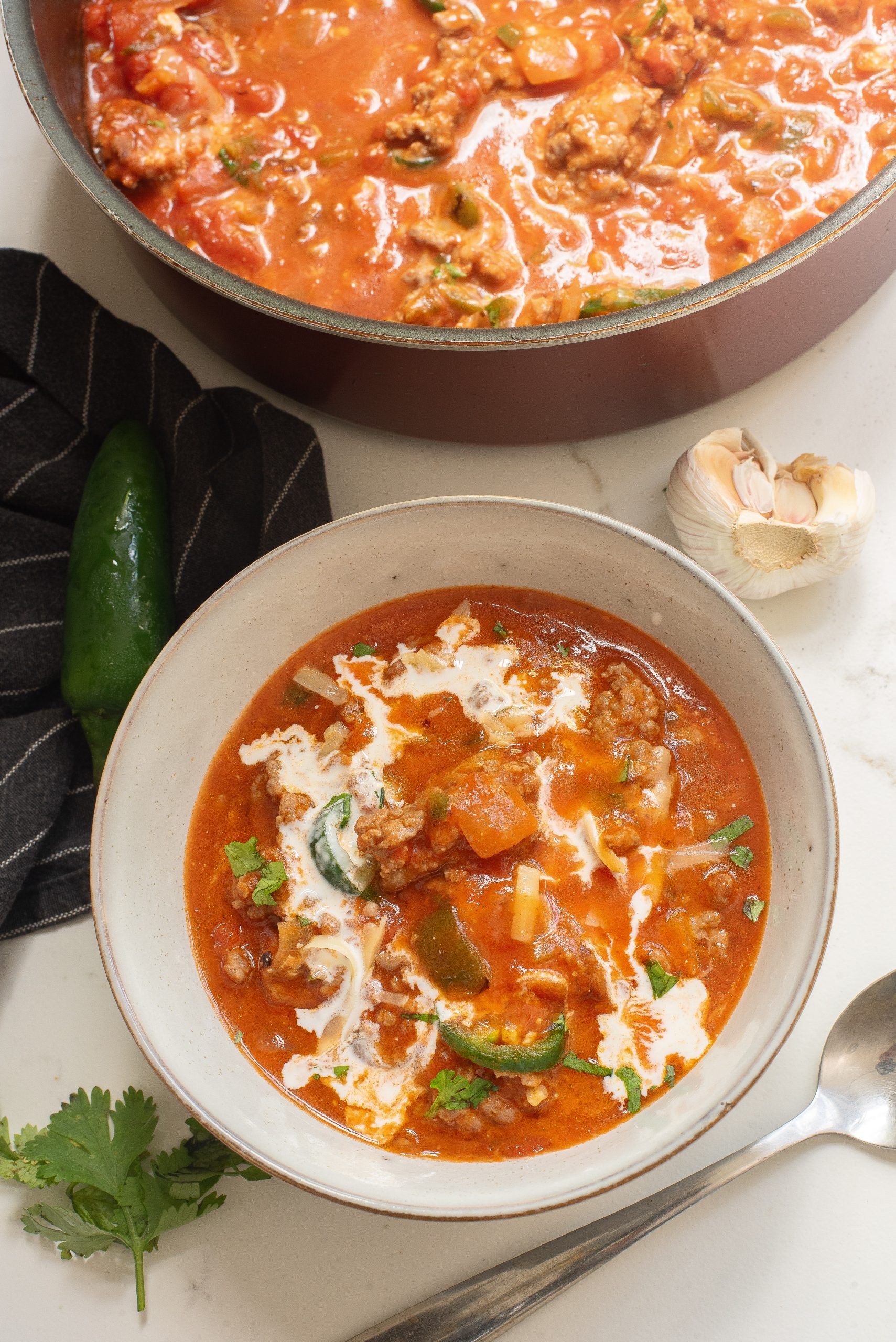 A bowl of spicy soup with sausage, peppers, and cream sits on a table next to a pot of soup, a jalapeño, cloves of garlic, and cilantro.