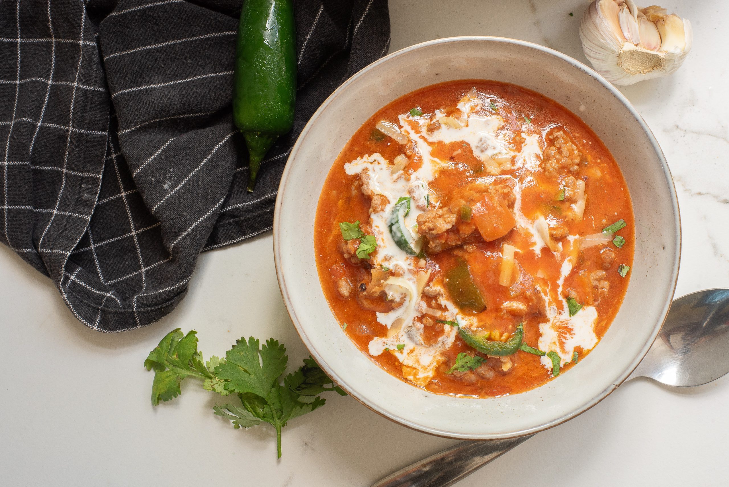 Bowl of red soup with meat, vegetables, and cream swirl on top. Garnished with sliced green chili and cilantro. Black cloth, jalapeño, garlic, and spoon beside.