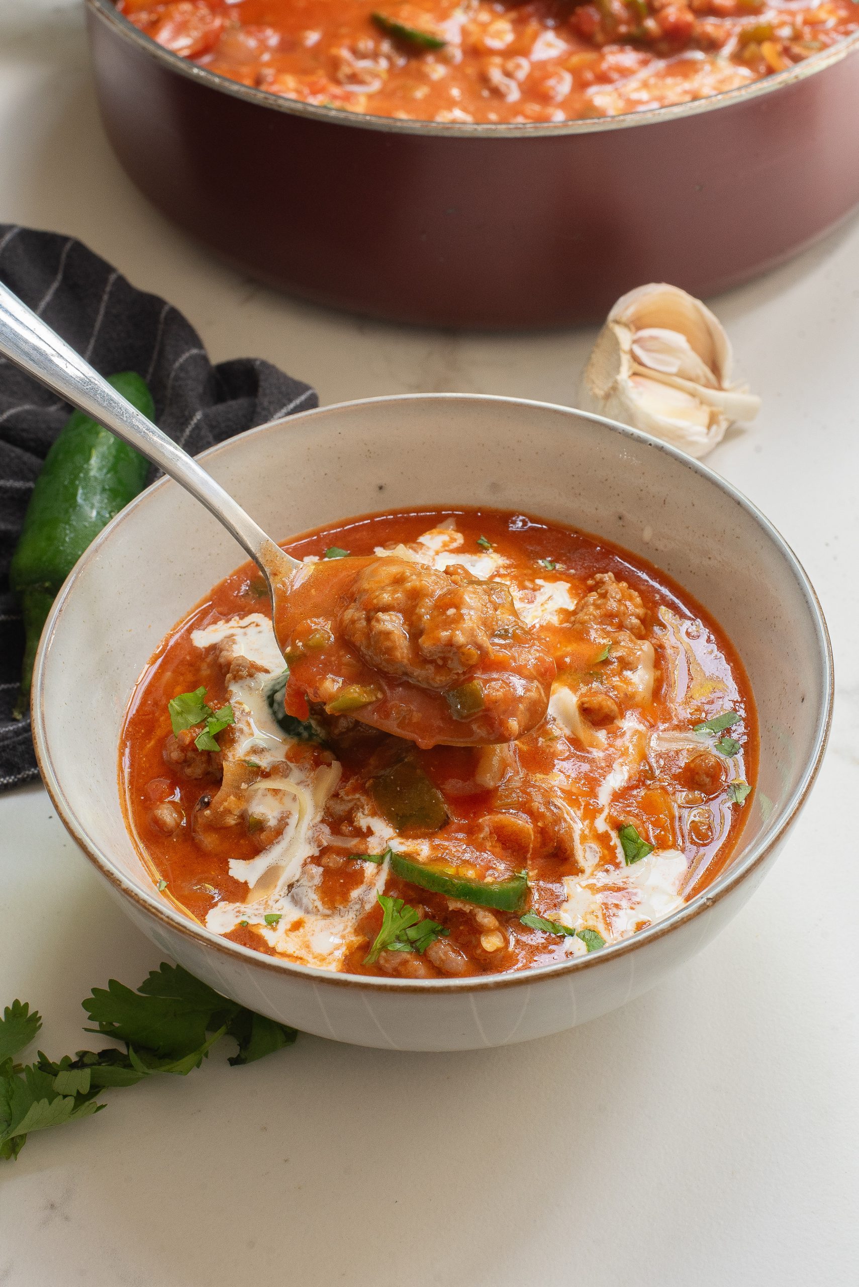 Bowl of tomato-based soup with vegetables and meat, topped with a swirl of cream. A spoon is dipping into the bowl. Brown pot and garlic are visible in the background.