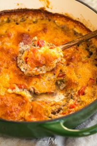 A close-up of a baked dish with melted cheese, ground meat, and tomatoes, being scooped with a spoon from a green pot.