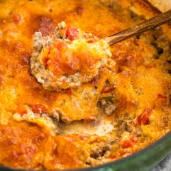 A close-up of a baked dish with melted cheese, ground meat, and tomatoes, being scooped with a spoon from a green pot.