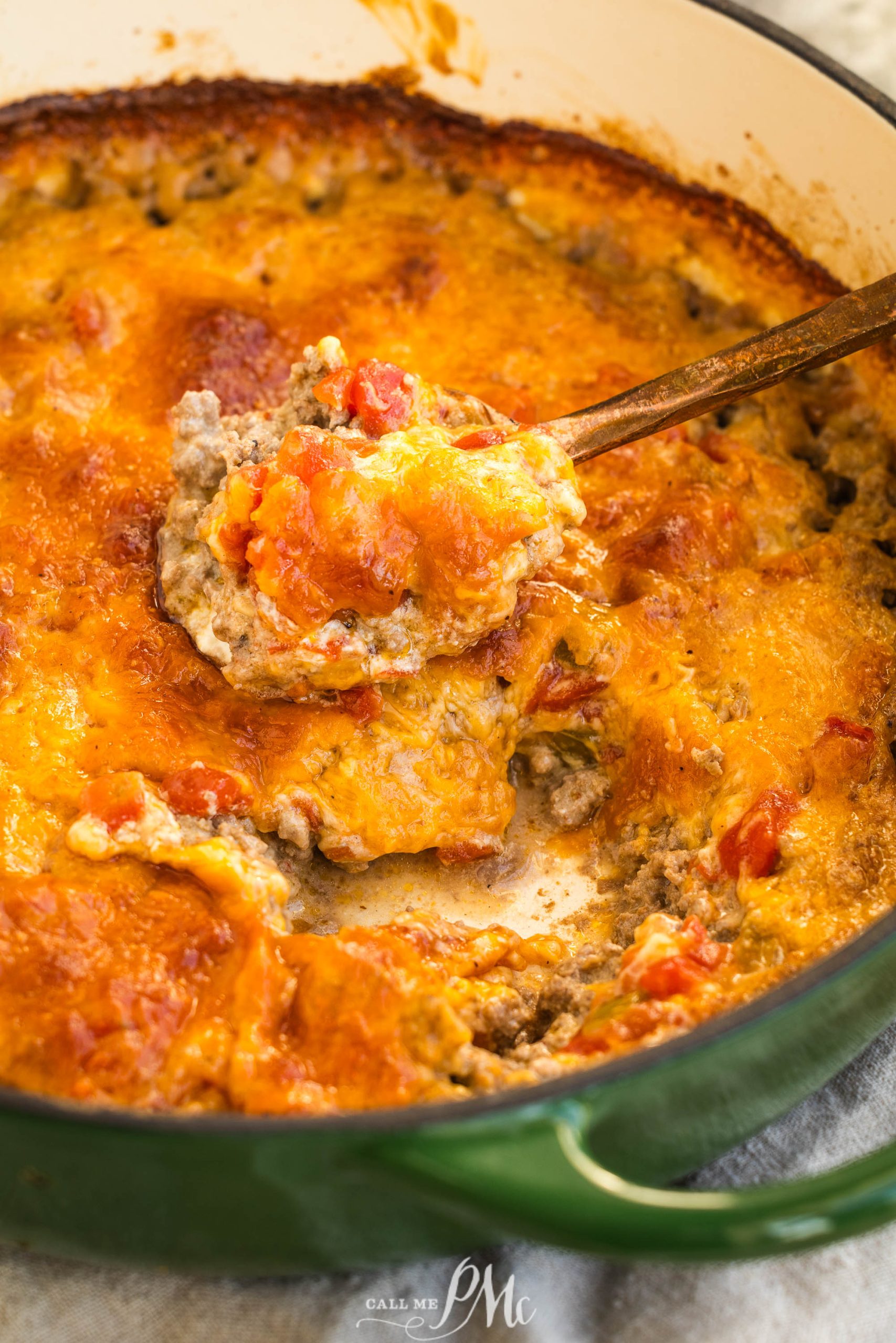 A close-up of Keto hamburger casserole with melted cheese, ground meat, and tomatoes, being scooped with a spoon from a green pot.