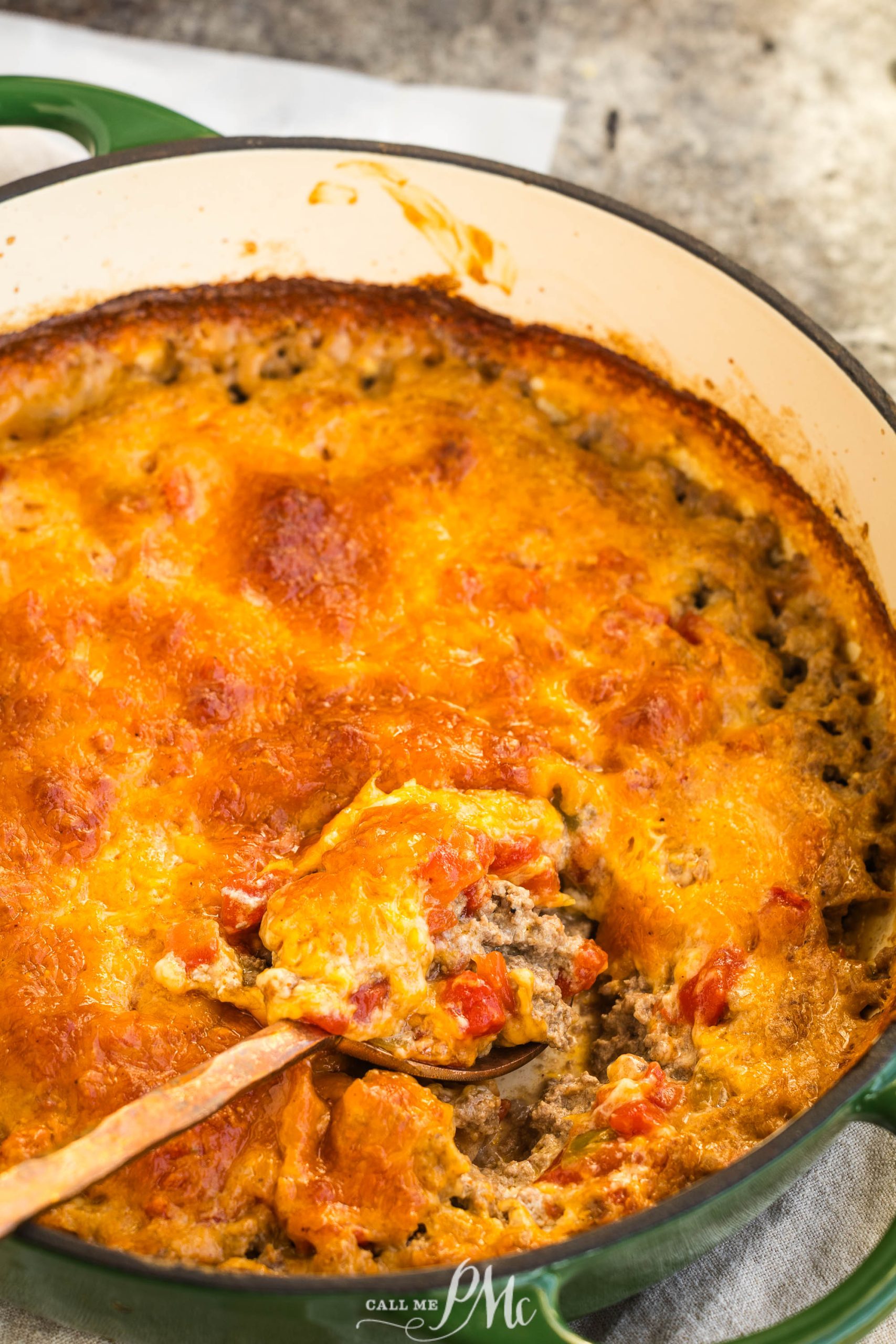 A keto hamburger casserole baked to perfection, featuring a golden-brown cheesy top with visible ground meat and vegetables nestled in a round green dish. A wooden spoon rests inside.