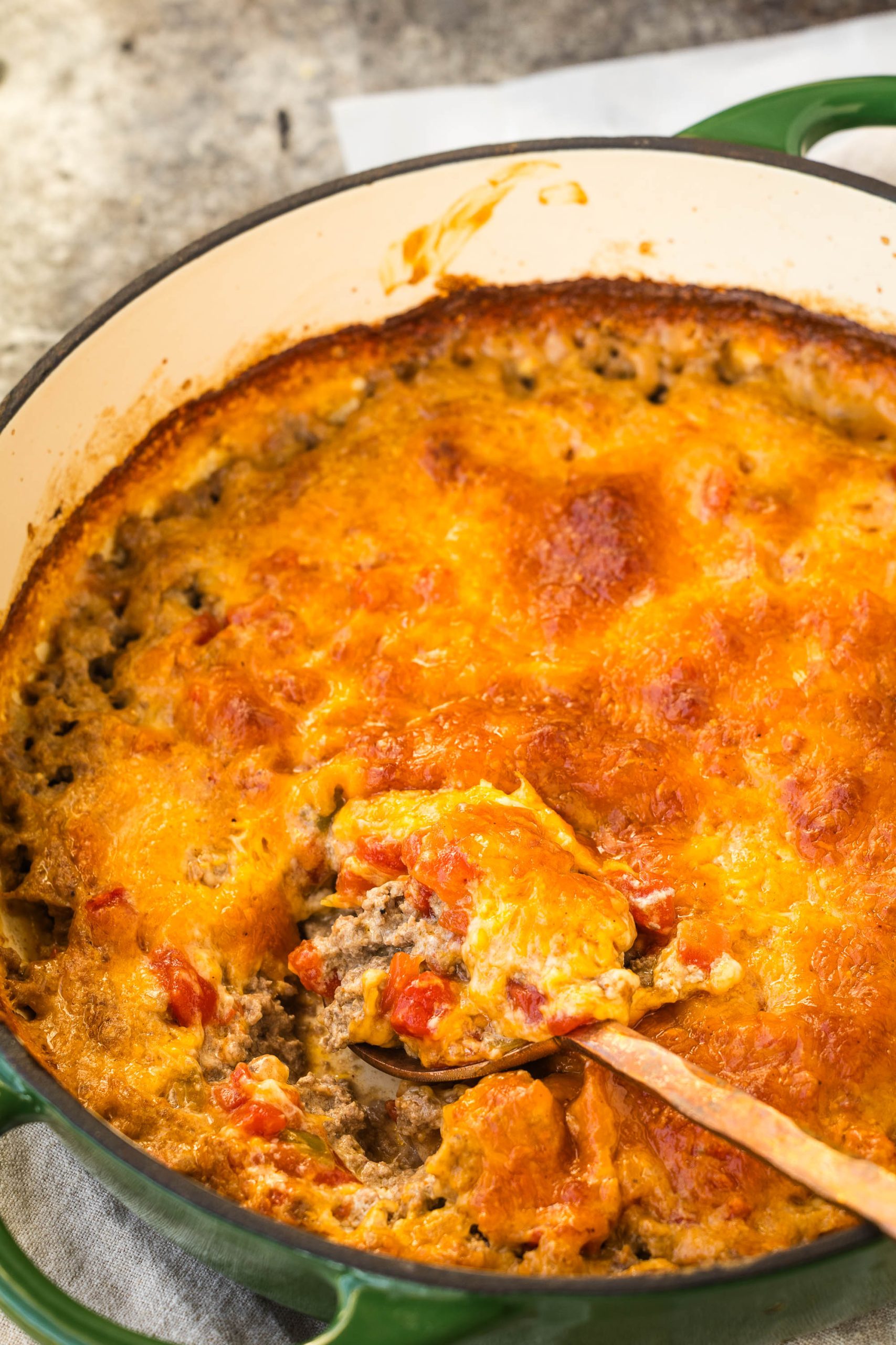 A baked casserole in a green dish, topped with a golden-brown layer of melted cheese and visible chunks of tomato. A wooden spoon is scooping into the dish.