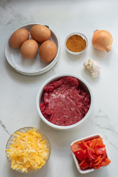 Ingredients on a countertop include eggs, seasoning, onion, garlic, raw beef, shredded cheese, and chopped red bell peppers.