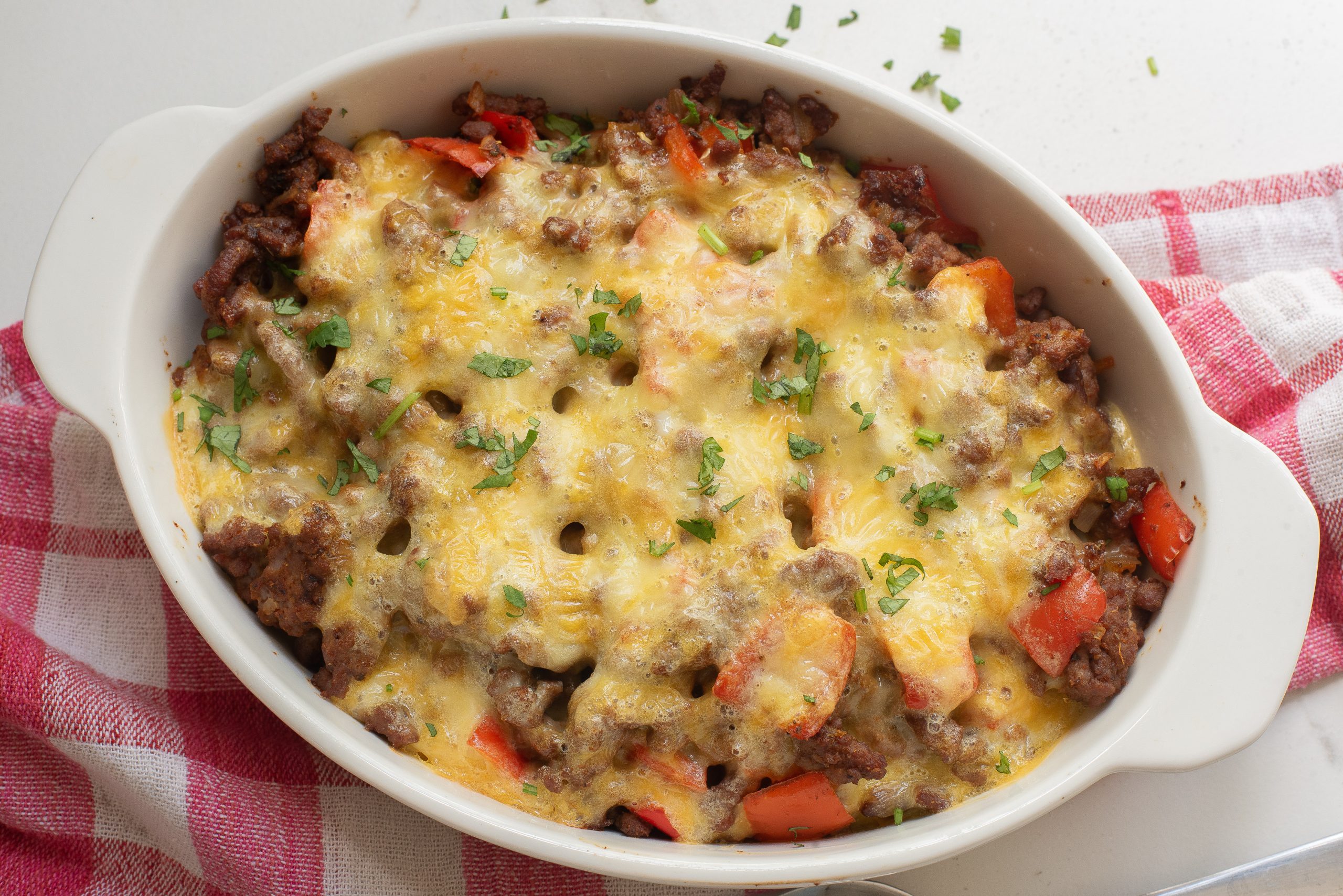 Keto Taco Casserole with melted cheese, ground meat, and diced red bell peppers in a white dish, garnished with chopped herbs, placed on a red and white checkered cloth.