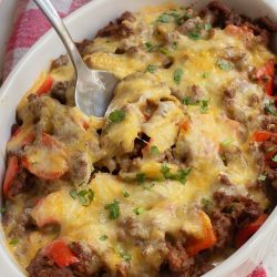 Cheesy baked casserole with ground meat, red peppers, and herbs in a white dish, served with a spoon on a red and white checkered cloth.