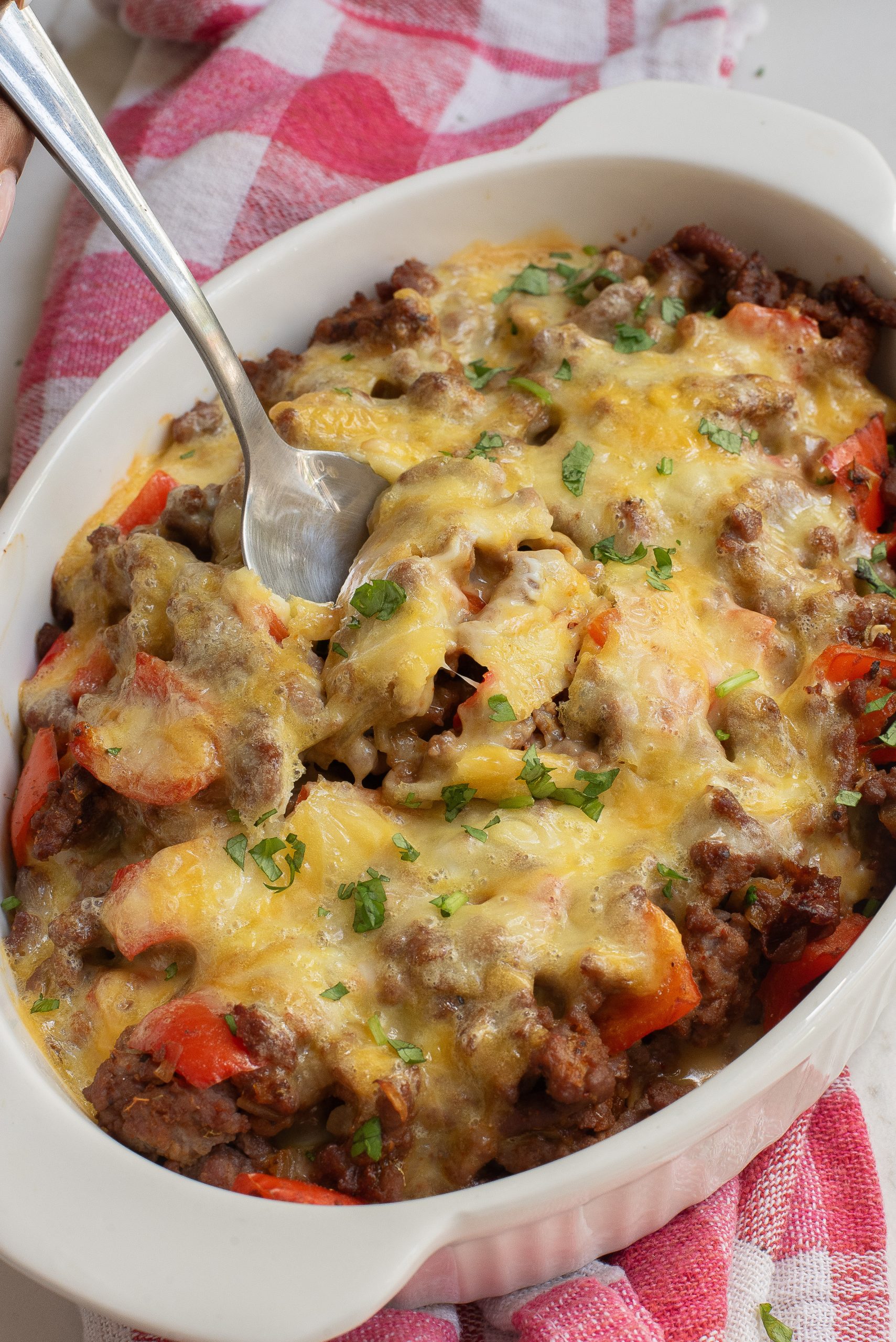 Cheesy baked casserole with ground meat, red peppers, and herbs in a white dish, served with a spoon on a red and white checkered cloth. It's an easy low carb recipes.