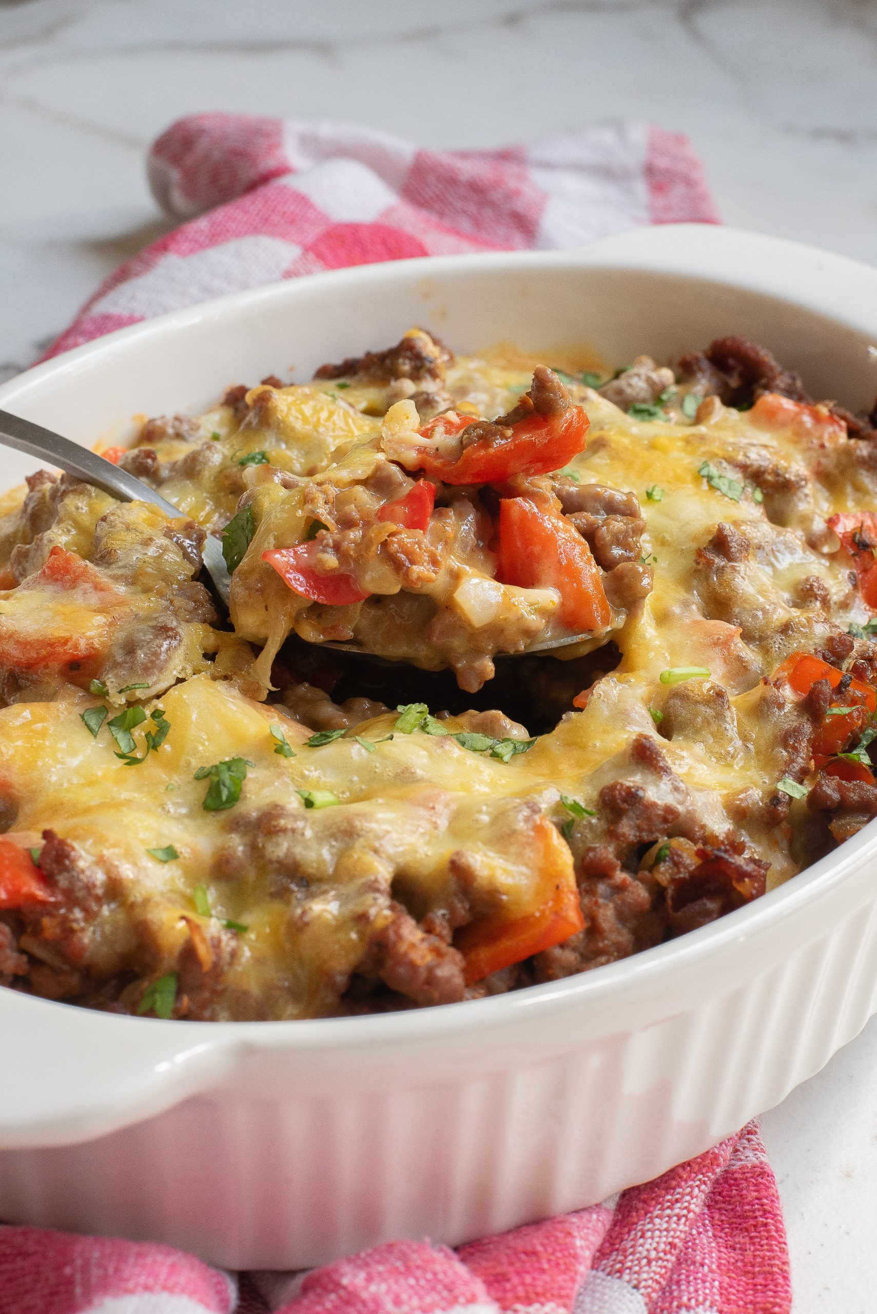 A cheesy casserole with ground beef, chopped red peppers, and herbs in a white dish on a red and white checkered cloth.