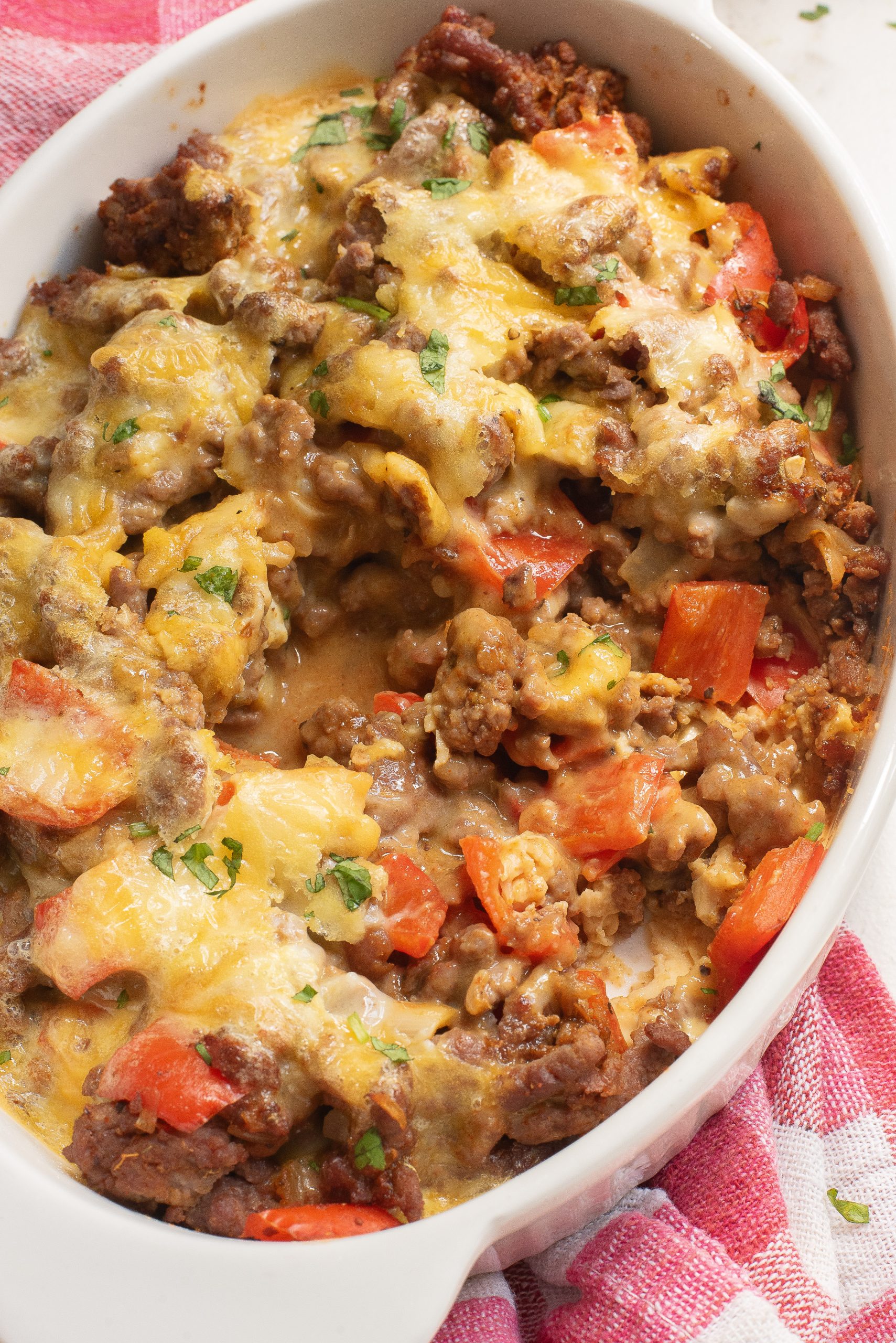 A casserole dish filled with baked ground beef, melted cheese, and chopped red peppers, garnished with herbs.