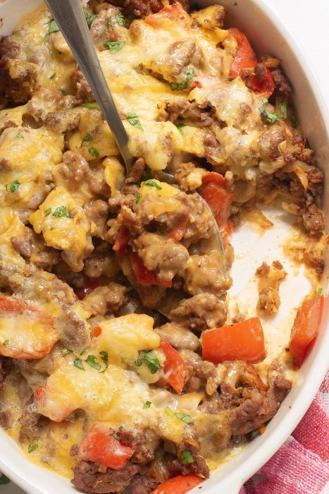 A Keto Taco Casserole with ground meat, melted cheese, red bell peppers, and green herbs in a white dish. A spoon is scooping a portion.