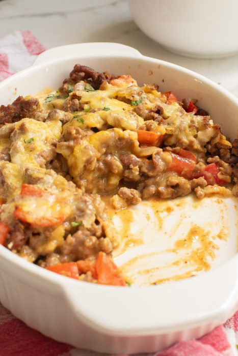 A white baking dish filled with a cheese-topped casserole that includes visible chunks of meat and red bell peppers. A portion is scooped out, revealing the ingredients and sauce.