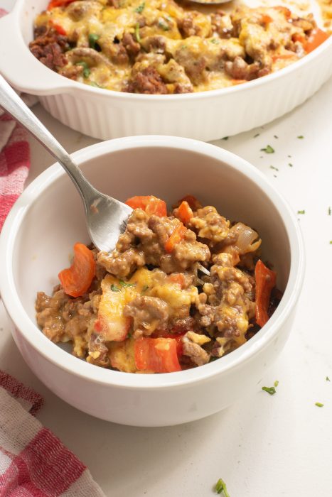 A white bowl with a serving of Keto Taco Casserole. A spoon rests in the bowl. Another dish with more of the same meal is partially visible in the background.