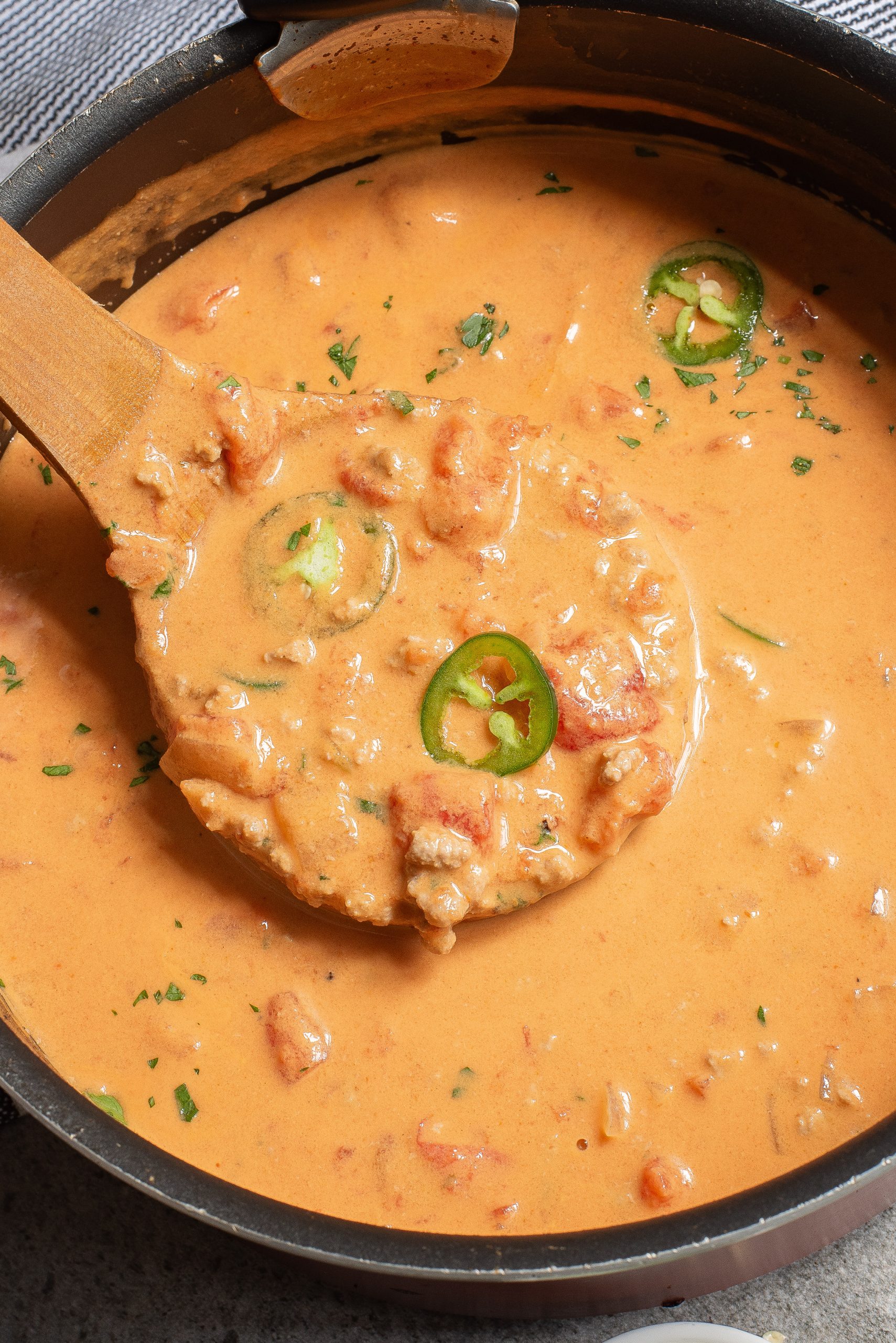 A wooden spoon lifts creamy queso dip from a pot, showing chunks of tomato and slices of jalapeño in easy low carb recipes.