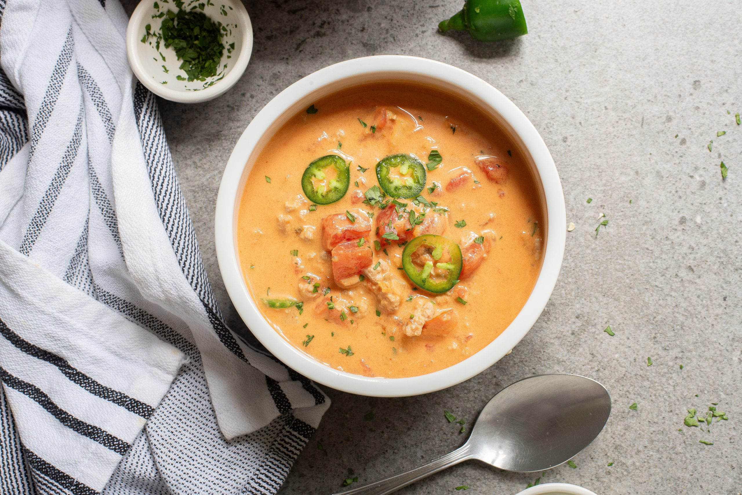 A bowl of soup garnished with sliced jalapeños and herbs, beside a spoon and striped napkin on a gray surface.