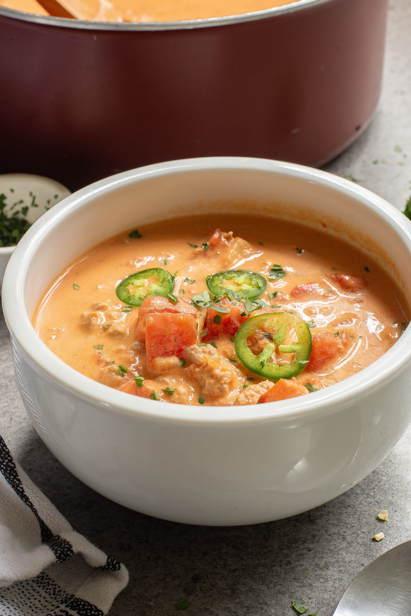 A bowl of Keto Taco Soup with chunks of tomato, ground meat, and sliced jalapeños, garnished with herbs. A pot and a dish of chopped herbs are in the background.