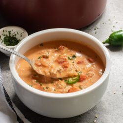 A bowl of creamy tomato soup with chunks of tomatoes and herbs, next to a pot. A spoon is in the bowl. Nearby, a jalapeño and small dish with herbs. A towel is partially visible.