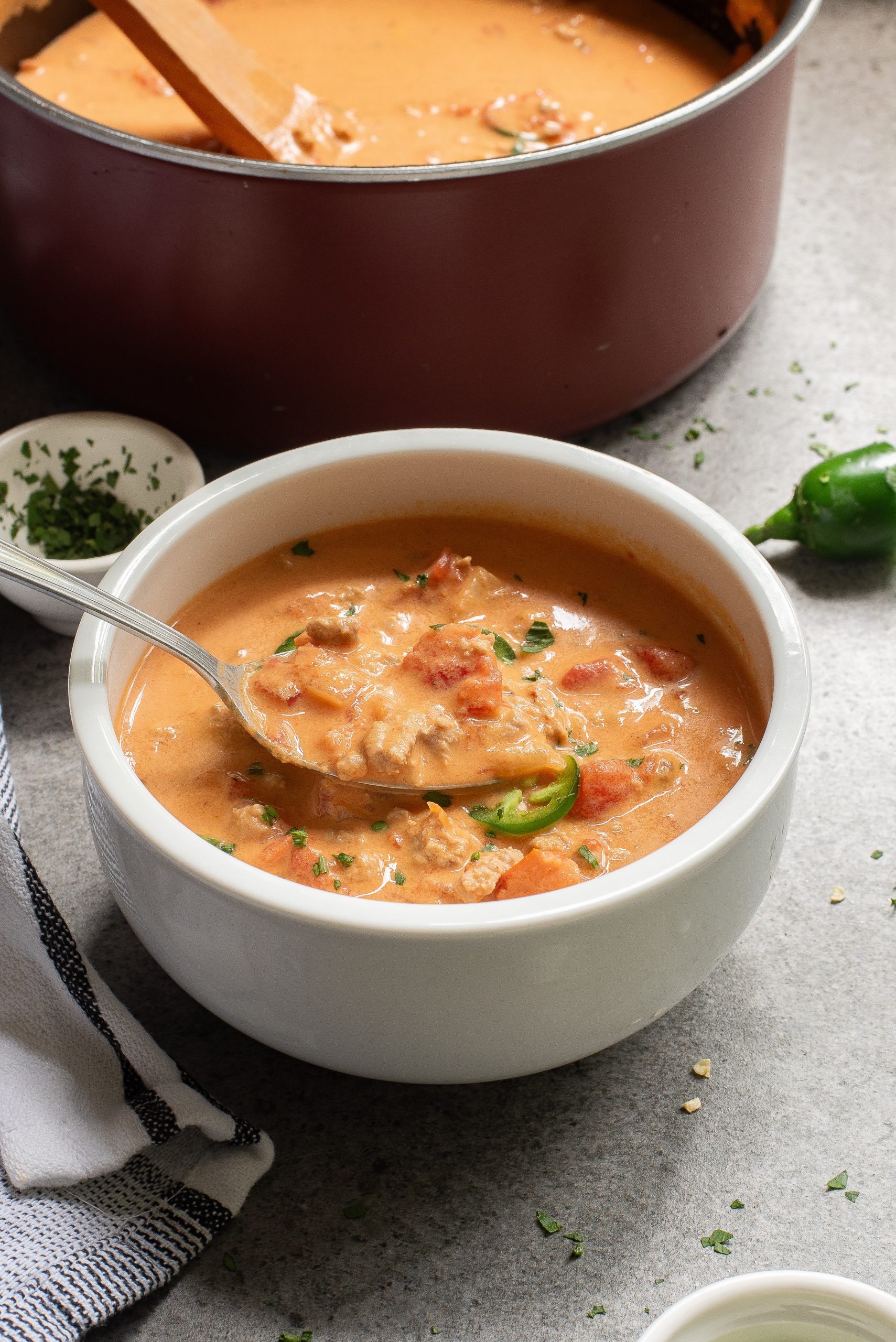 A bowl of Keto Taco Soup with chunks of tomatoes and herbs, next to a pot. A spoon is in the bowl. Nearby, a jalapeño and small dish with herbs. A towel is partially visible.