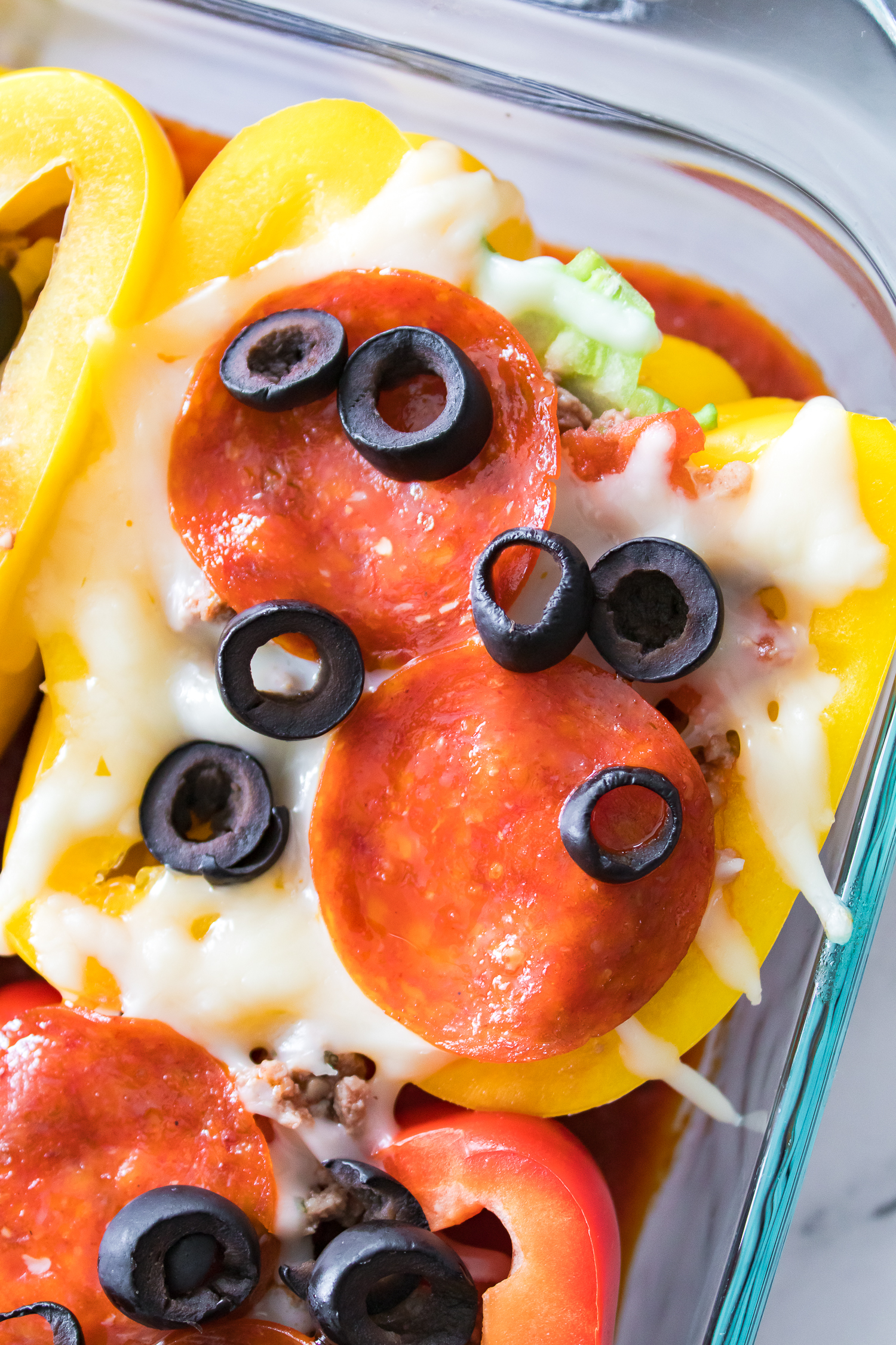 Close-up of a dish with yellow and red bell peppers, topped with melted cheese, pepperoni slices, black olives, and a tomato-based sauce in a glass dish.