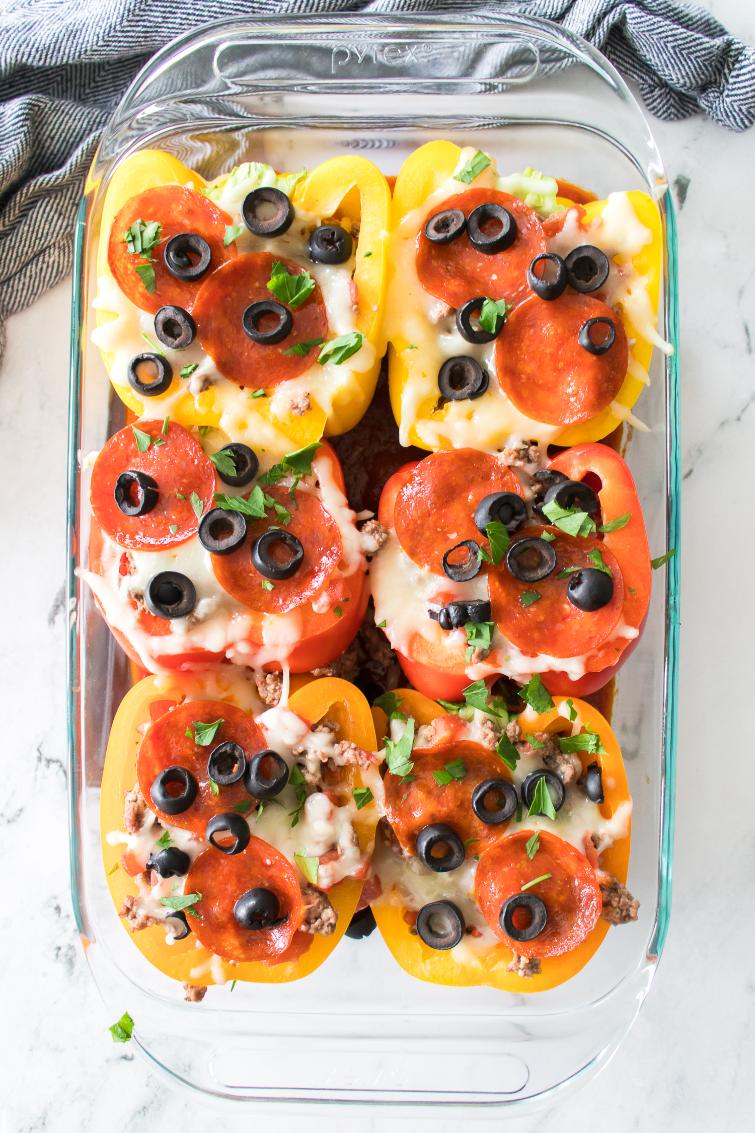 A glass dish with six stuffed bell peppers topped with melted cheese, pepperoni, black olives, and herbs. Peppers are colored red and yellow.