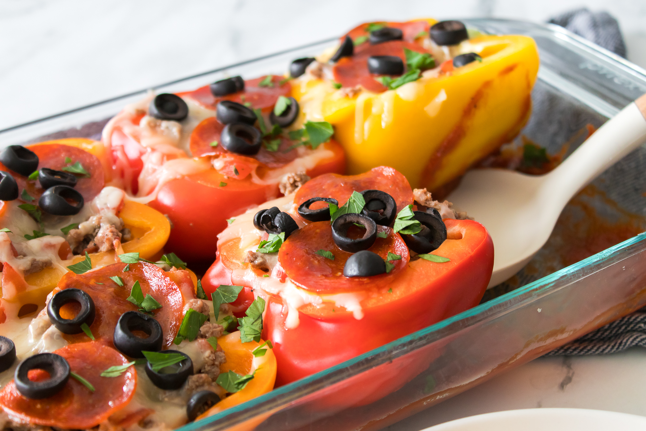 A glass dish containing stuffed bell peppers with ground meat, tomato sauce, melted cheese, sliced black olives, and chopped parsley.