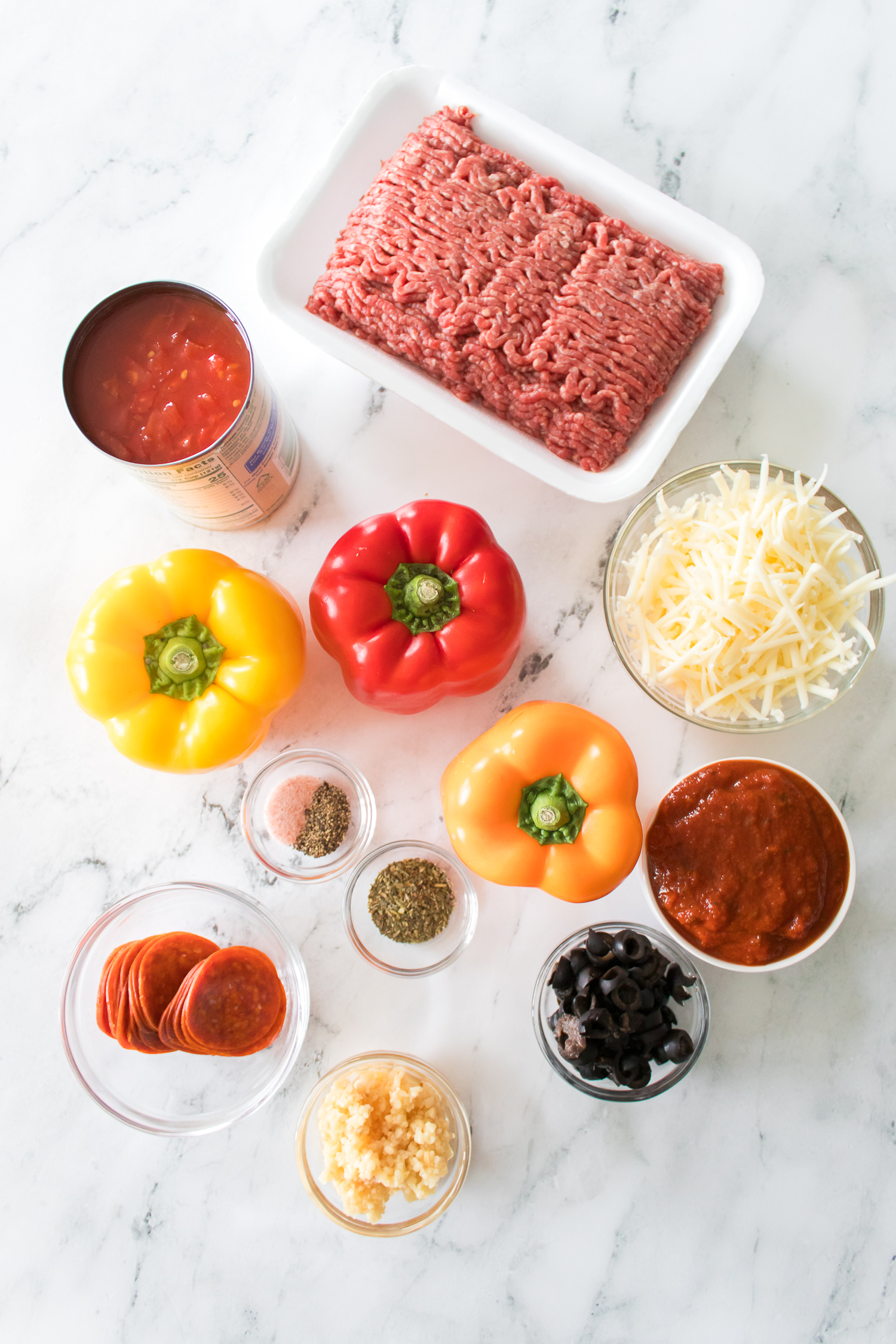 Ingredients for stuffed peppers on a marble surface include ground beef, shredded cheese, bell peppers, tomato sauce, diced tomatoes, pepperoni, olives, spices, and minced garlic.