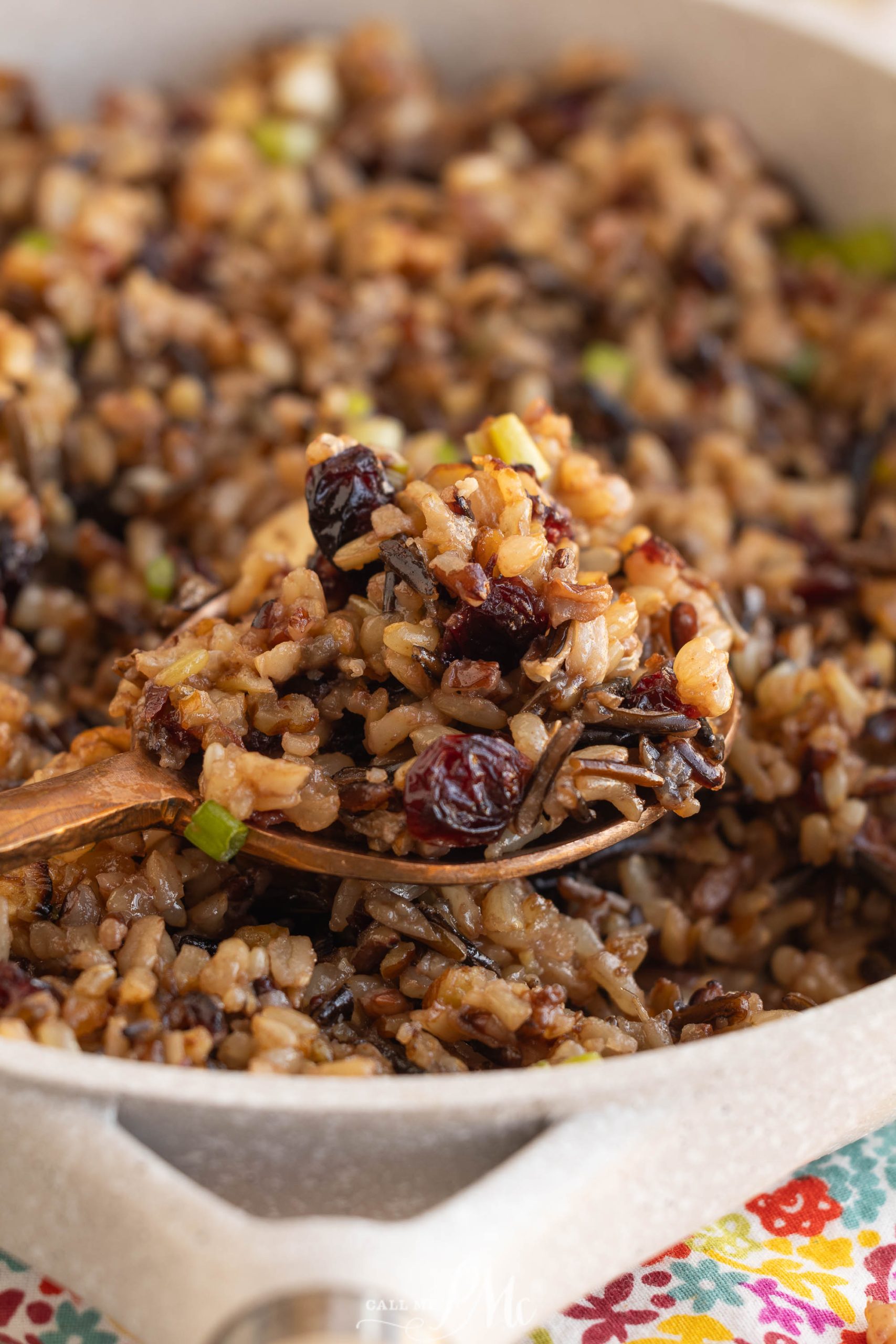 A spoon is lifting a portion of wild rice dish mixed with cranberries and green onions from a pan.