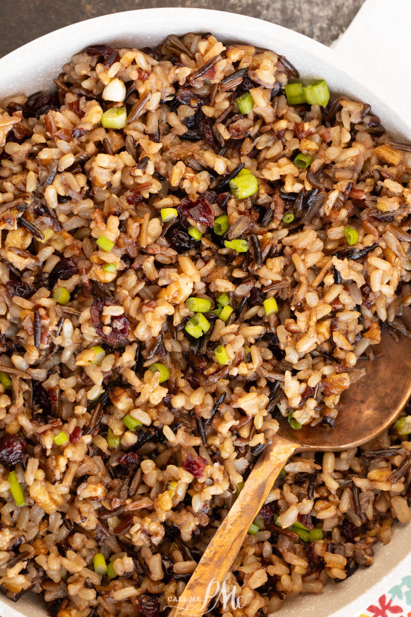 Close-up of a bowl filled with food mixed with cranberries, nuts, and green onions, accompanied by a serving spoon.