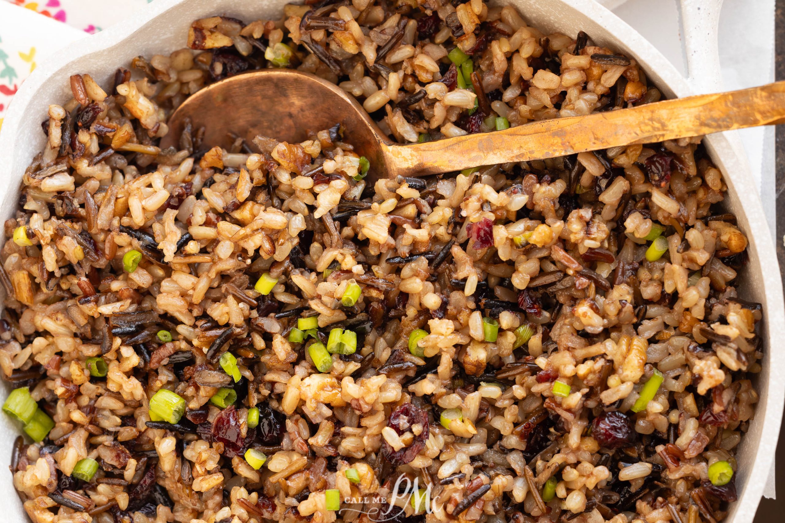 A close-up of a bowl filled with cooked wild rice mixed with chopped green onions and cranberries, with a wooden spoon resting on top.