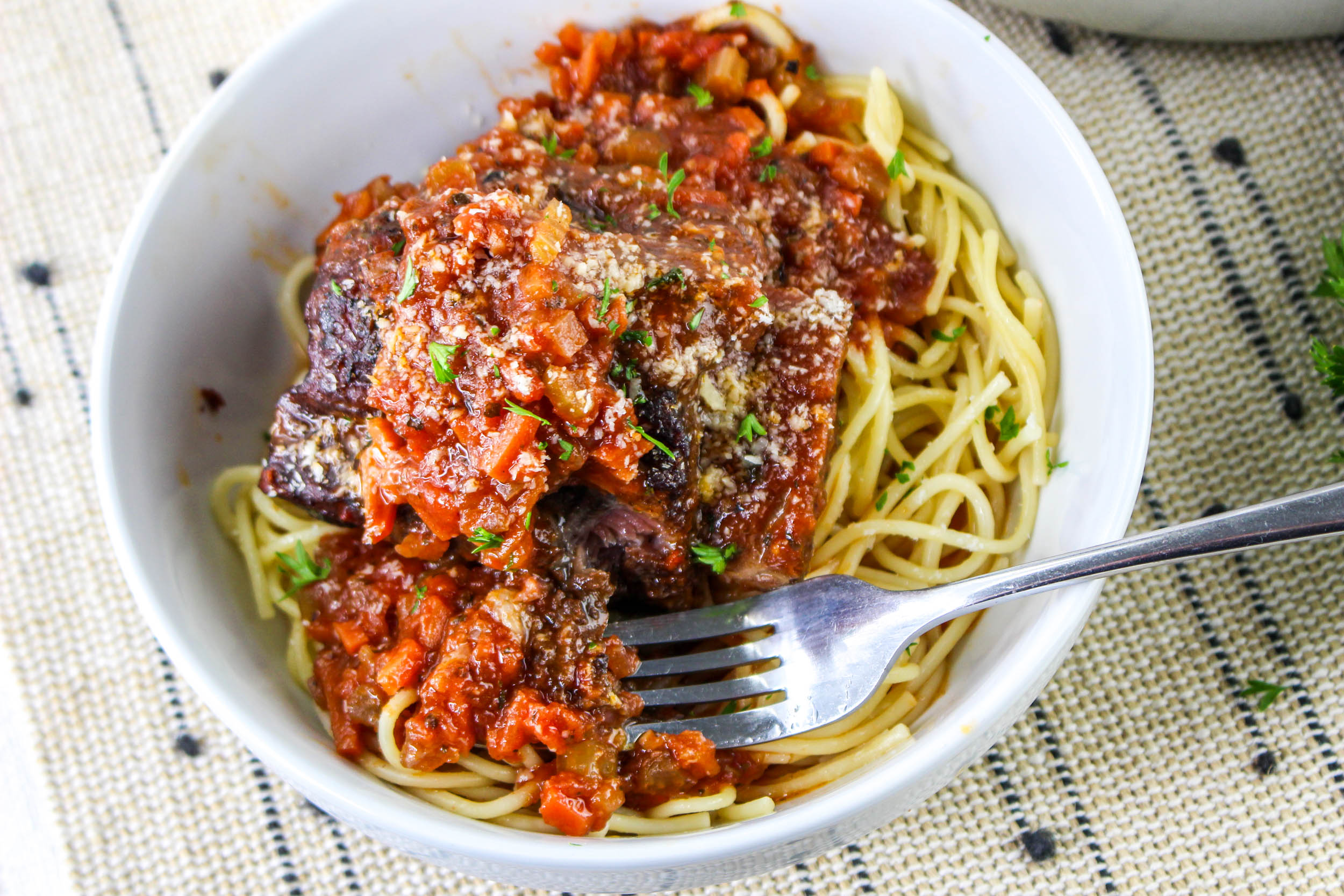 Bowl of spaghetti topped with tomato sauce and a beef piece, garnished with herbs and grated cheese; a fork is resting inside.