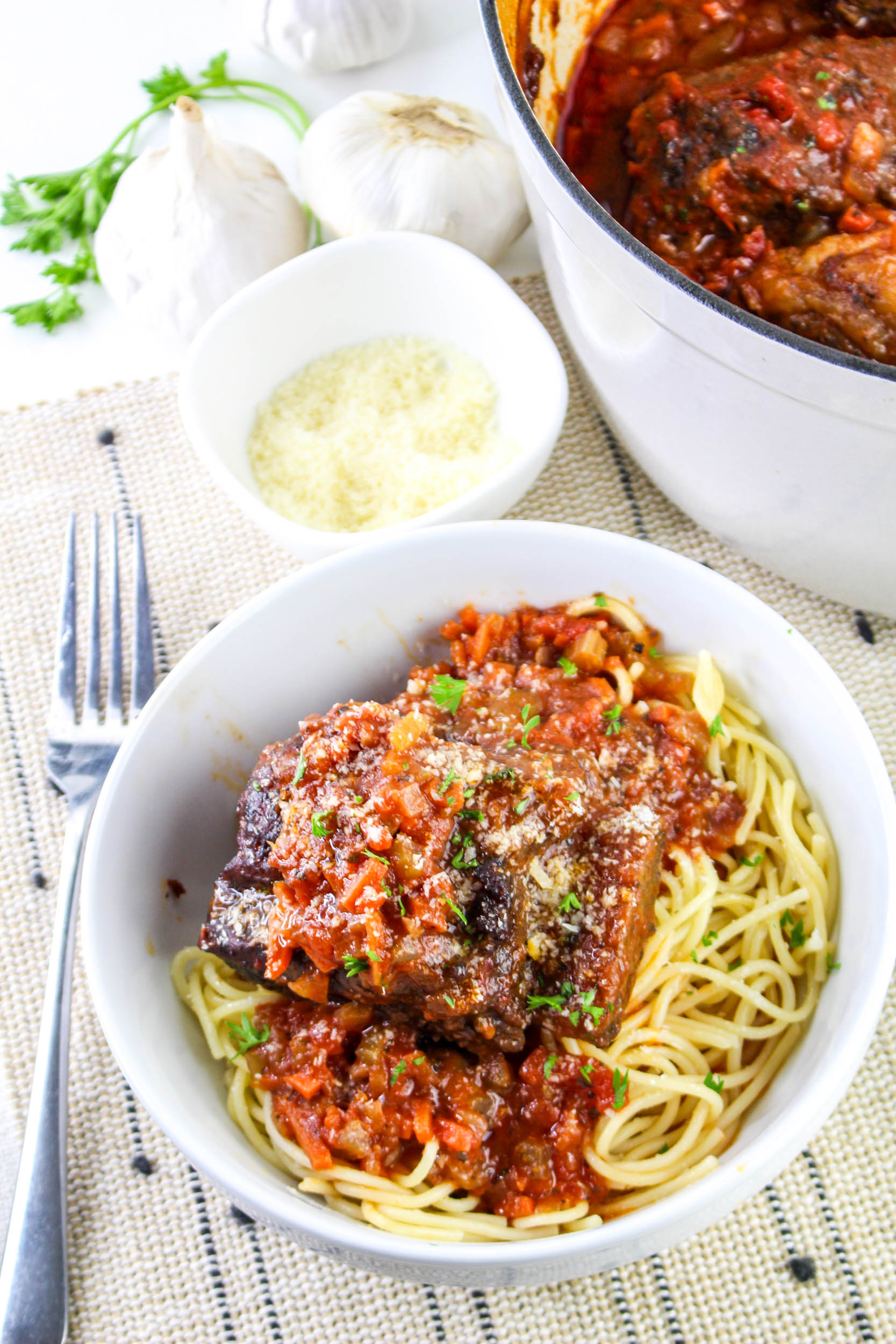 A bowl of spaghetti with beef ribs in tomato sauce, topped with grated cheese and herbs. A pot of sauce, a small bowl of cheese, garlic, and a fork are nearby.