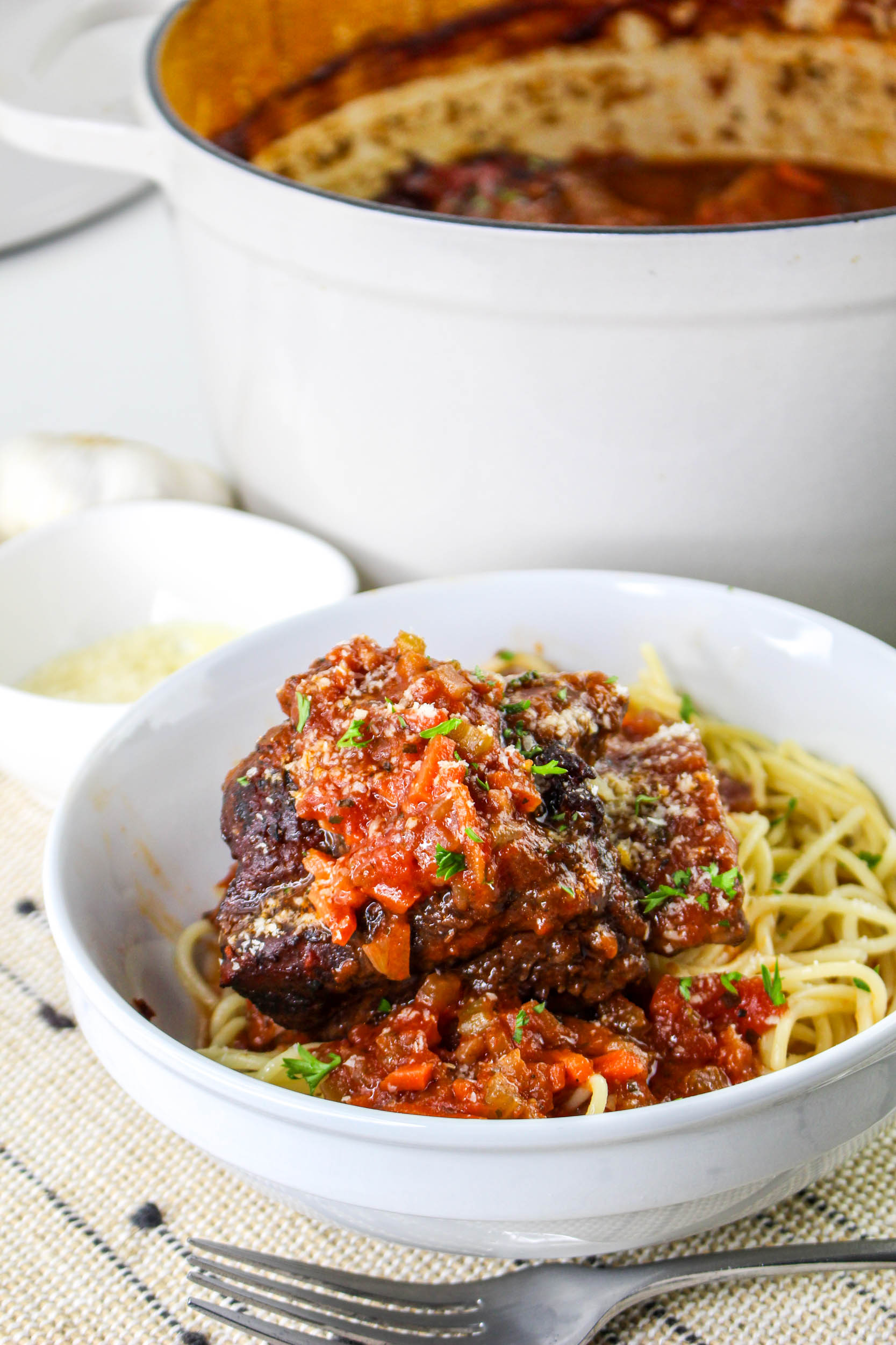 A bowl of spaghetti topped with beef oxtail stew and tomato sauce, garnished with parsley. A pot and bowl of grated cheese are in the background on a beige cloth.