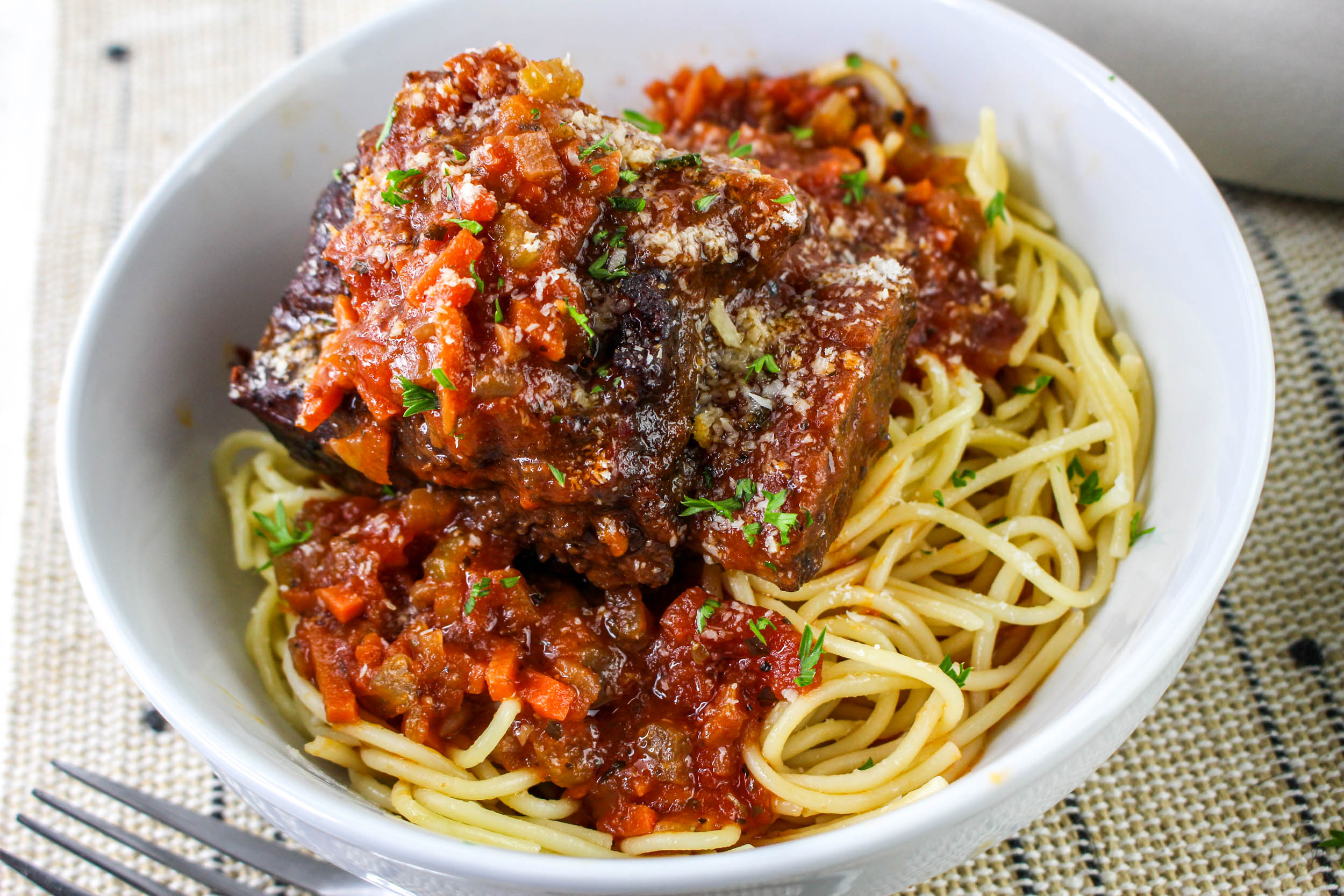 Pasta topped with a rich tomato sauce and braised beef in a white bowl on a patterned placemat.