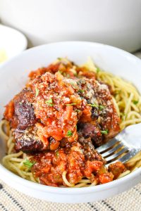 Bowl of spaghetti topped with braised beef in tomato sauce, garnished with herbs and grated cheese. A fork rests beside the pasta.