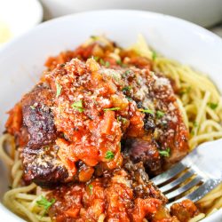 Bowl of spaghetti topped with braised beef in tomato sauce, garnished with herbs and grated cheese. A fork rests beside the pasta.