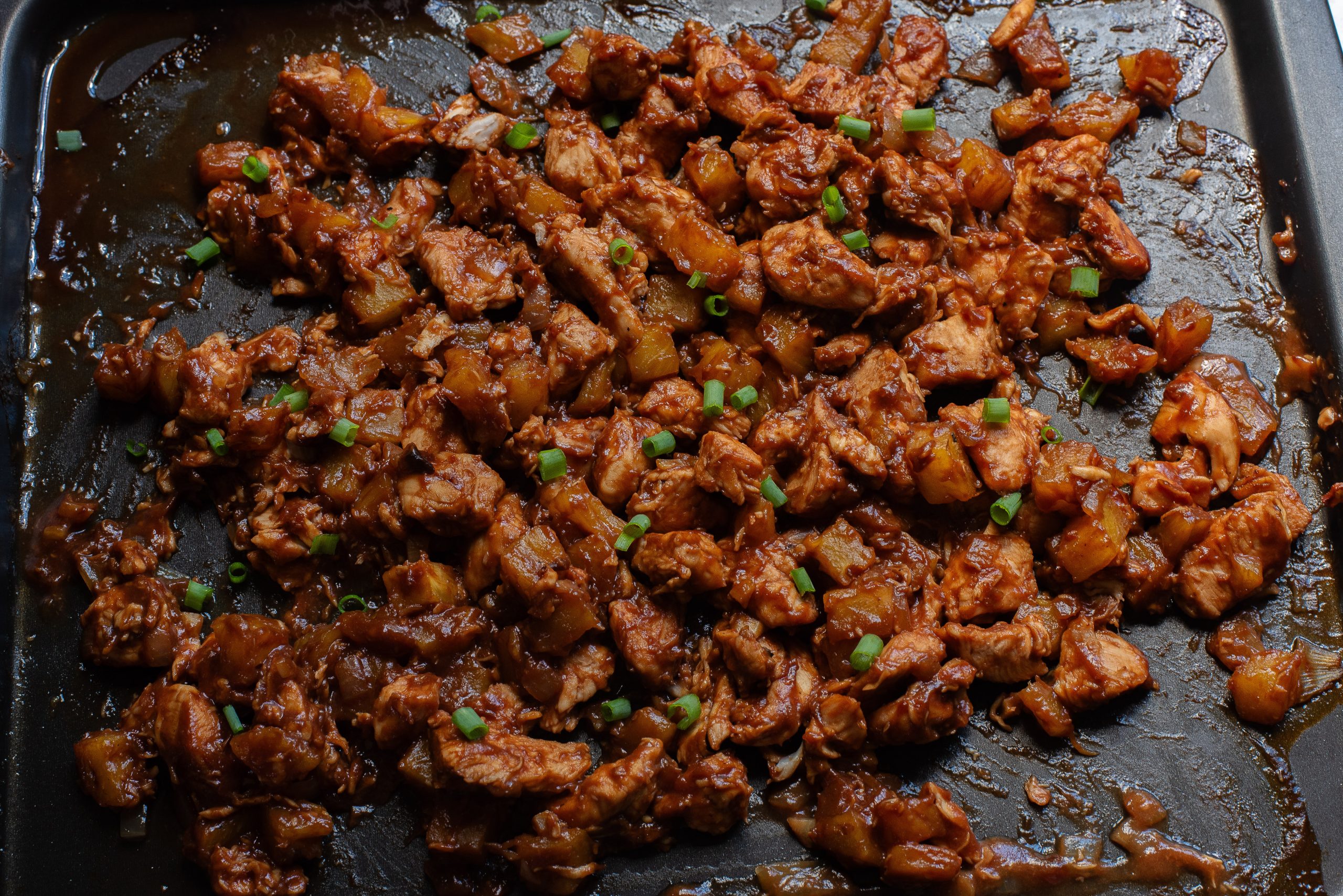 Cooked diced meat with sauce and green onion garnishes on a black baking tray.