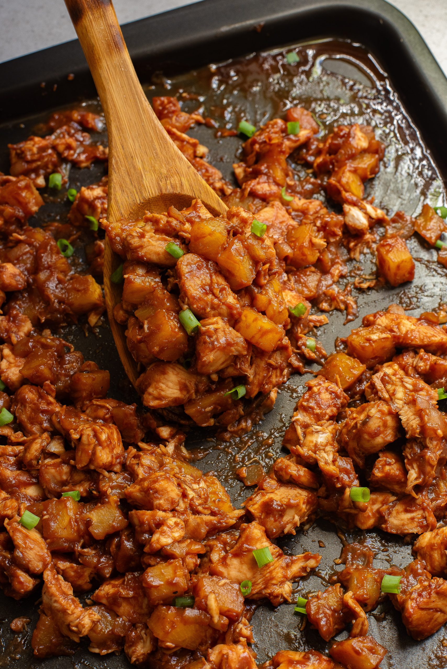 Cooked chicken and pineapple pieces on a baking tray with green onions, mixed with sauce, and a wooden spoon scooping some.