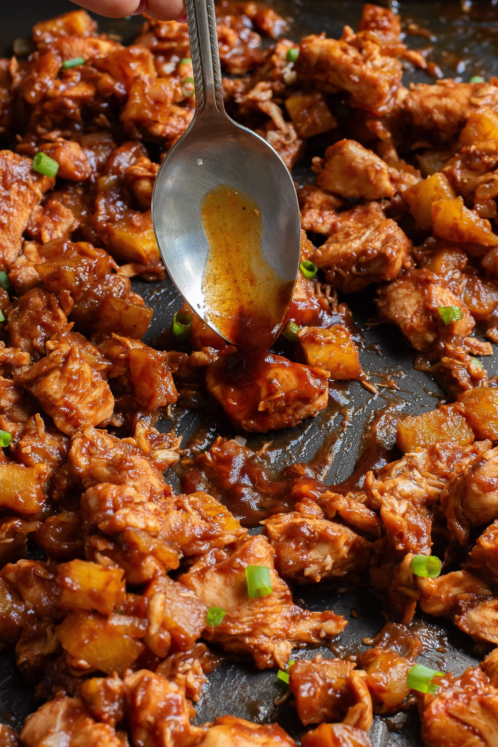 A spoon drizzles sauce over cooked chicken pieces with pineapple chunks and green onions on a dark surface.