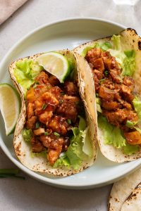 Two tacos with grilled meat, lettuce, and sauce, topped with green onions, served on a plate with lime wedges.