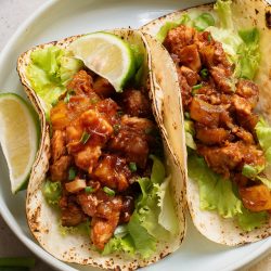 Two tacos with grilled meat, lettuce, and sauce, topped with green onions, served on a plate with lime wedges.