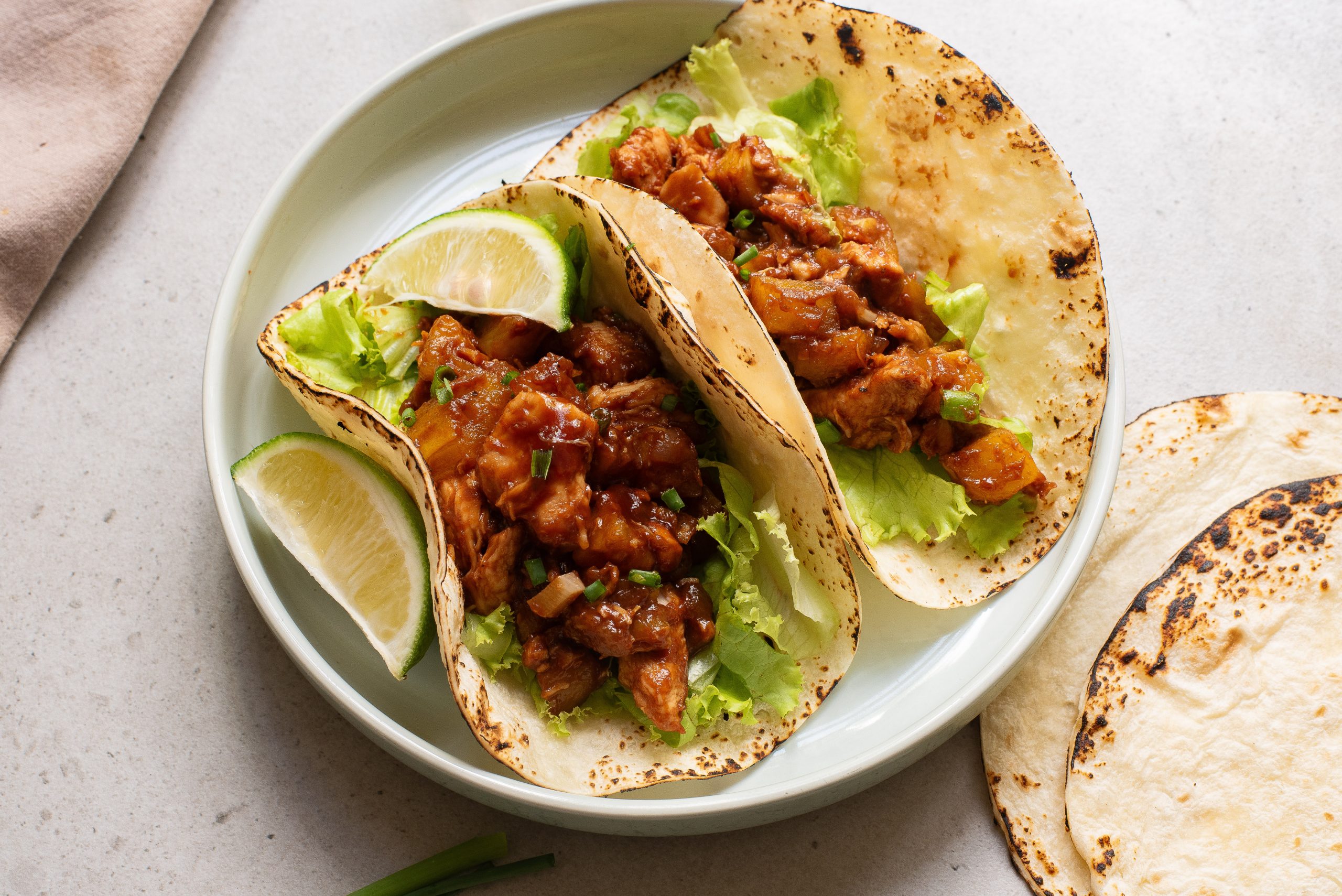 Two BBQ Chicken Sheet Pan Tacos filled with seasoned meat and lettuce, garnished with lime wedges, served on a white plate.