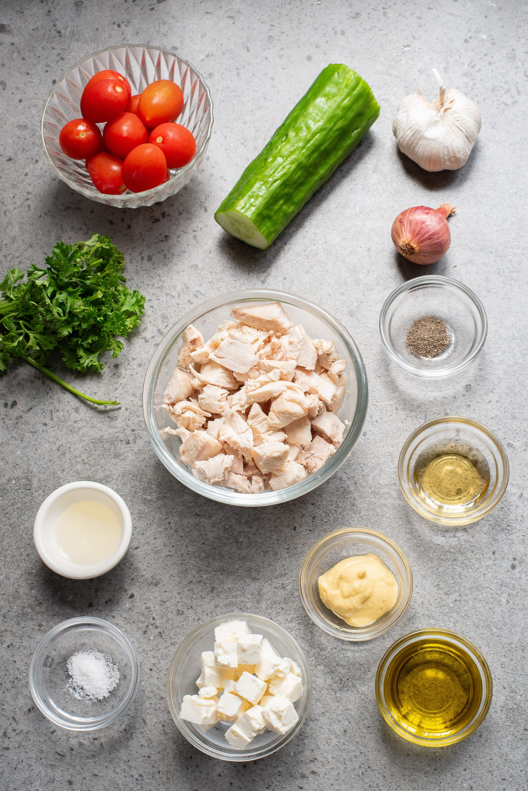 Ingredients for a salad laid out: cherry tomatoes, cucumber, garlic, shallot, parsley, shredded chicken, cheese, mustard, olive oil, vinegar, pepper, and salt on a gray surface.