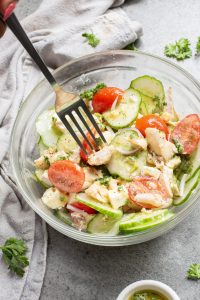 A fork is placed in a bowl of salad containing sliced cucumbers, cherry tomatoes, pieces of chicken, and parsley, on a gray surface with a cloth and parsley scattered around.