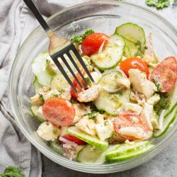 A fork is placed in a bowl of salad containing sliced cucumbers, cherry tomatoes, pieces of chicken, and parsley, on a gray surface with a cloth and parsley scattered around.