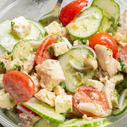 A clear bowl filled with a salad of cucumber slices, cherry tomatoes, diced chicken, feta cheese, and chopped herbs, with a fork.